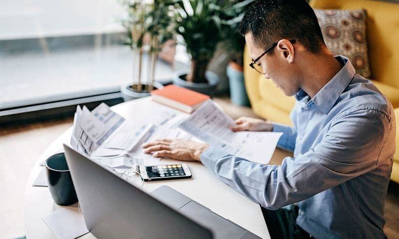 A developer reviewing paper bills with a calculator at a desk beside a laptop