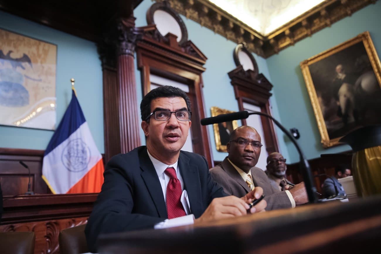 Inside a New York City Council hearing chamber: a council member speaks at the wooden dais with the NYC flag and classical decor behind, a microphone facing the speaker.