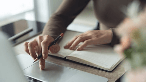 Real photo of a person writing in a notebook beside a laptop