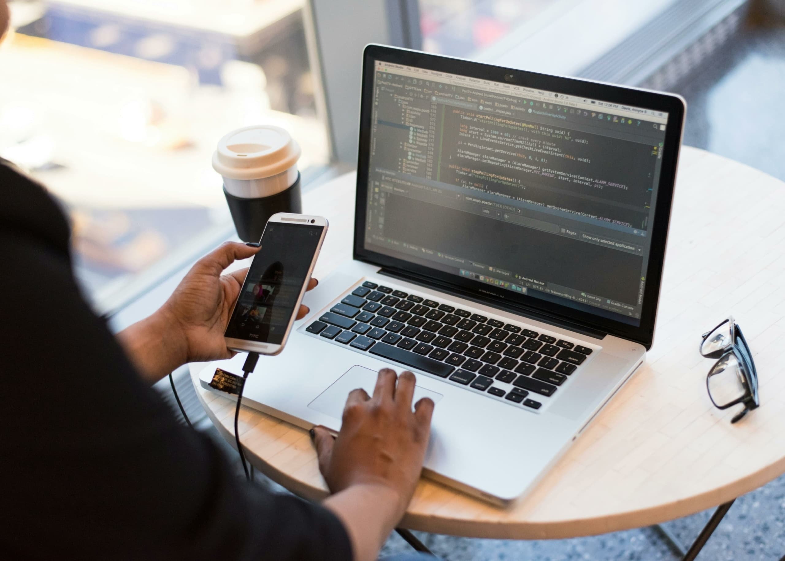 A real photograph of a person holding an Android phone beside a laptop showing Android Studio code; it is not an official Google announcement screenshot.