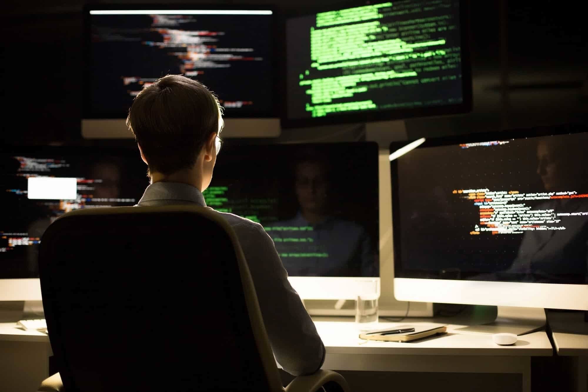 A developer seen from behind in a dark office, facing multiple screens showing code
