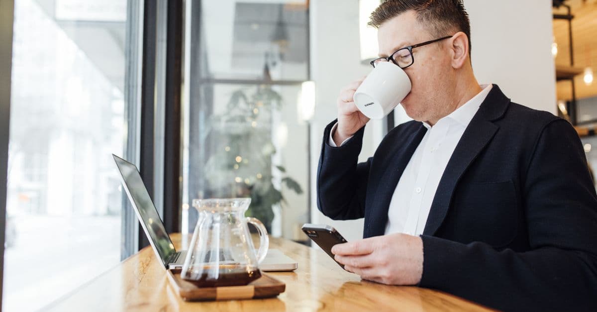 A man drinking coffee at a morning table while holding his phone, with a laptop in front of him, a real photo echoing Duet's morning-brief scene