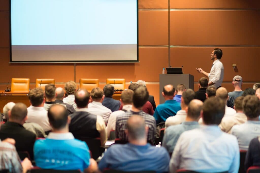 A real photograph of a presenter speaking to an audience in a hall, standing in for a live product demo delivered to real people.