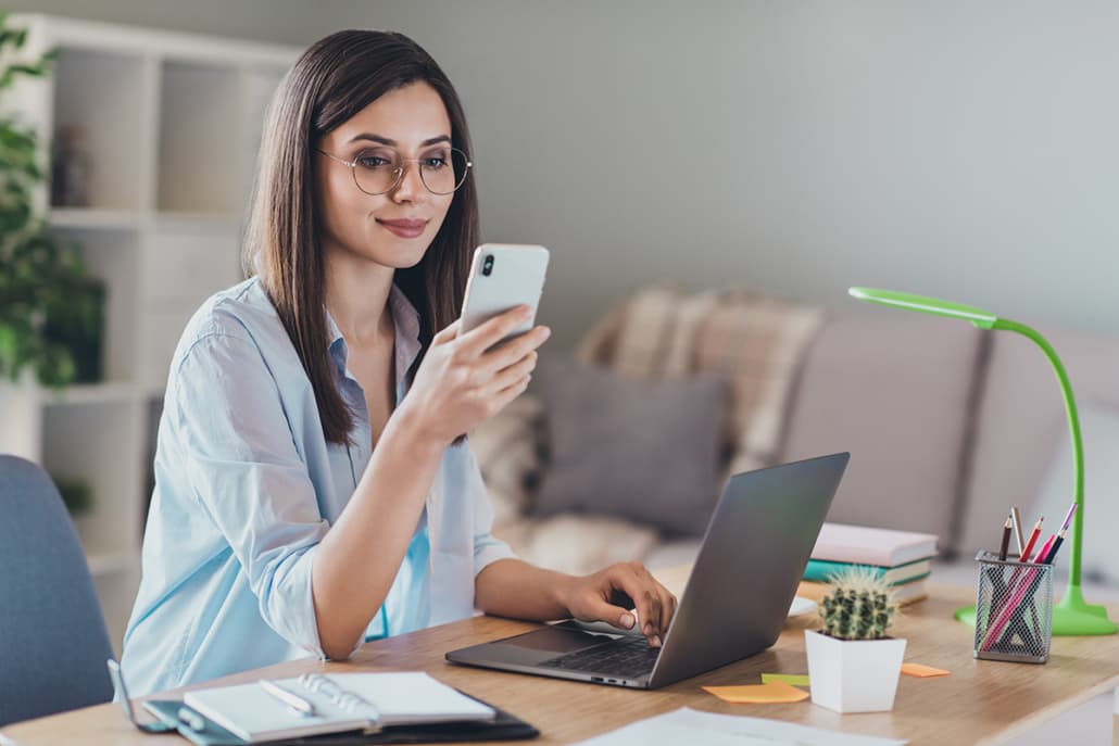 A real photo of a woman holding a phone while working on a laptop, illustrating multi-device login sessions