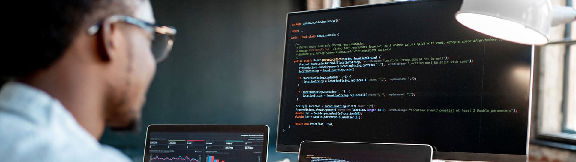 A developer at a desk looking at a large screen showing code, with a laptop beside it