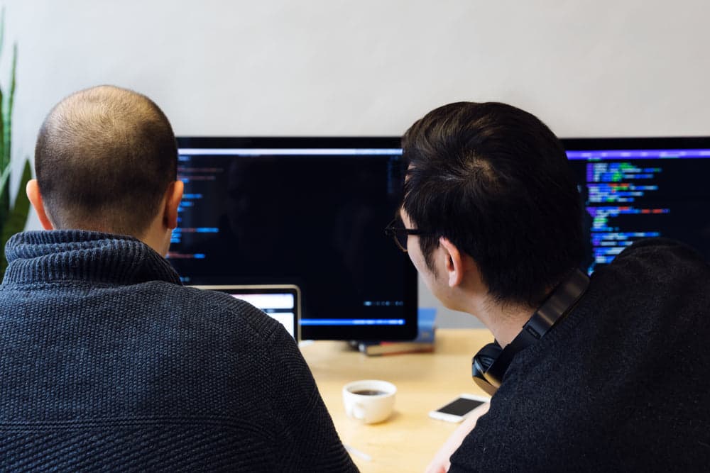 A real photograph of two developers sitting before screens showing code, reviewing together; it is not a screenshot of the GitHub Copilot product interface.