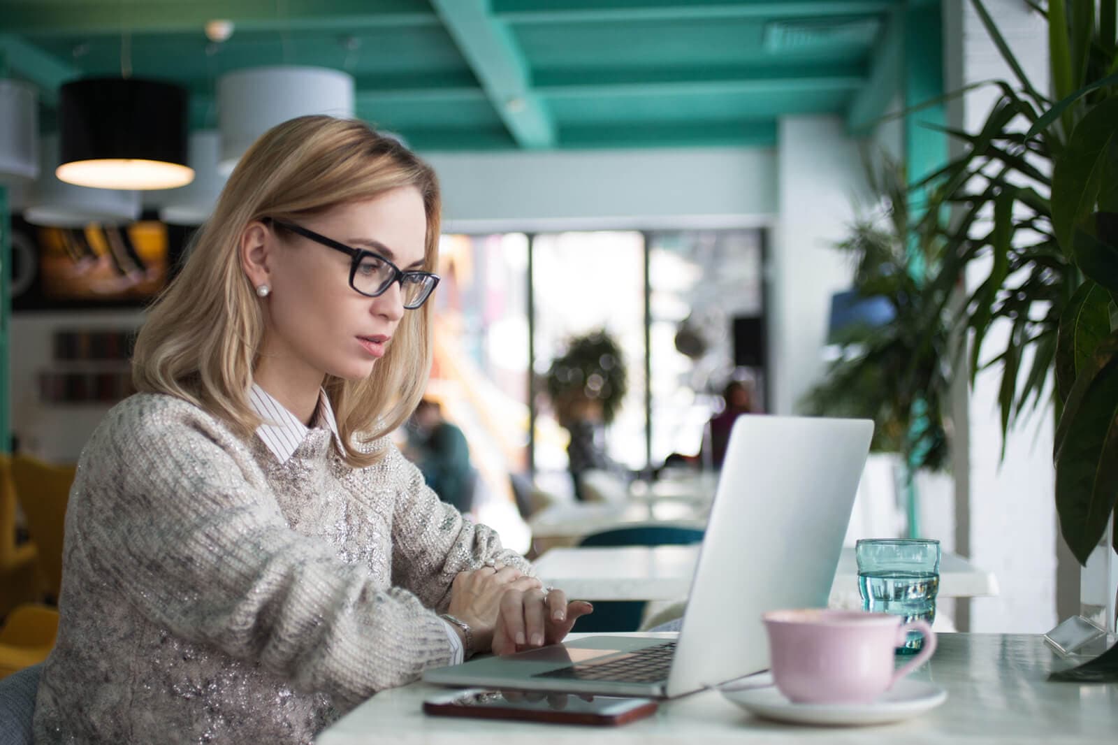 A woman with glasses using a laptop at a cafe table with a coffee cup beside her, trying out an app for the first time