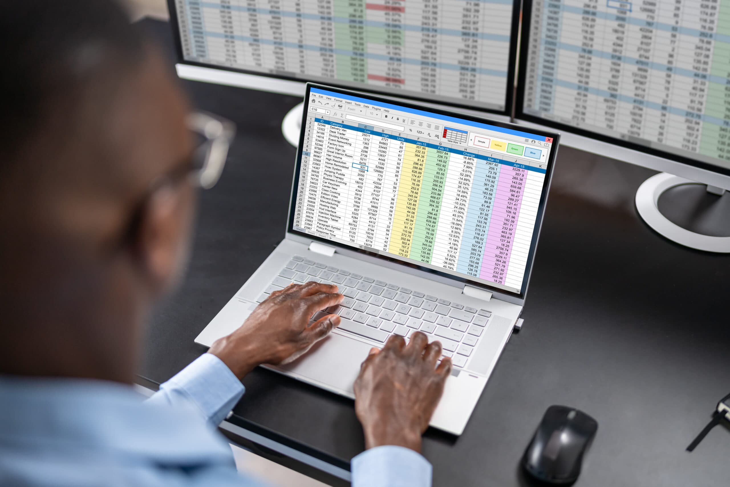 A man working at a laptop showing spreadsheets on screen, with two large monitors behind him also displaying spreadsheet data