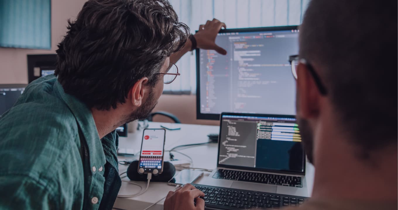 A real photo of two developers discussing code at a screen together; not a product screenshot