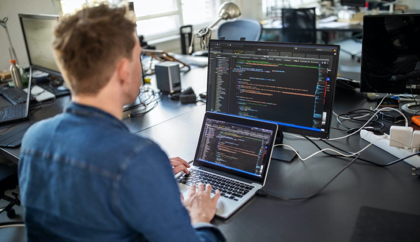 Cover: a real photo of a developer writing code on a laptop with an external monitor at a desk
