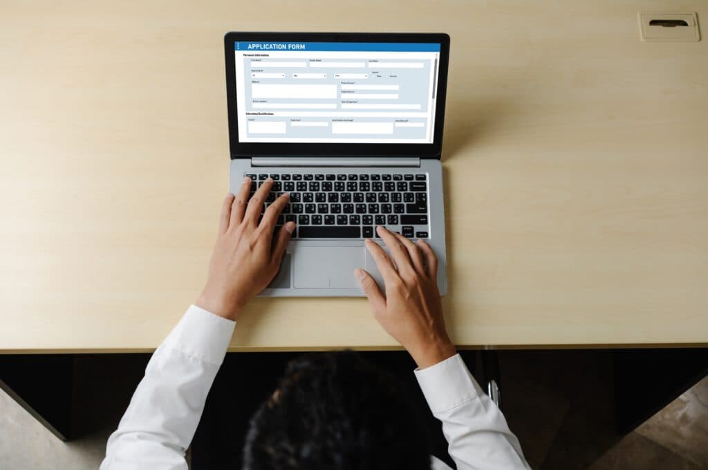A real photograph, viewed from above, of hands typing on a laptop showing an online application form; it is not a screenshot of a specific form product.