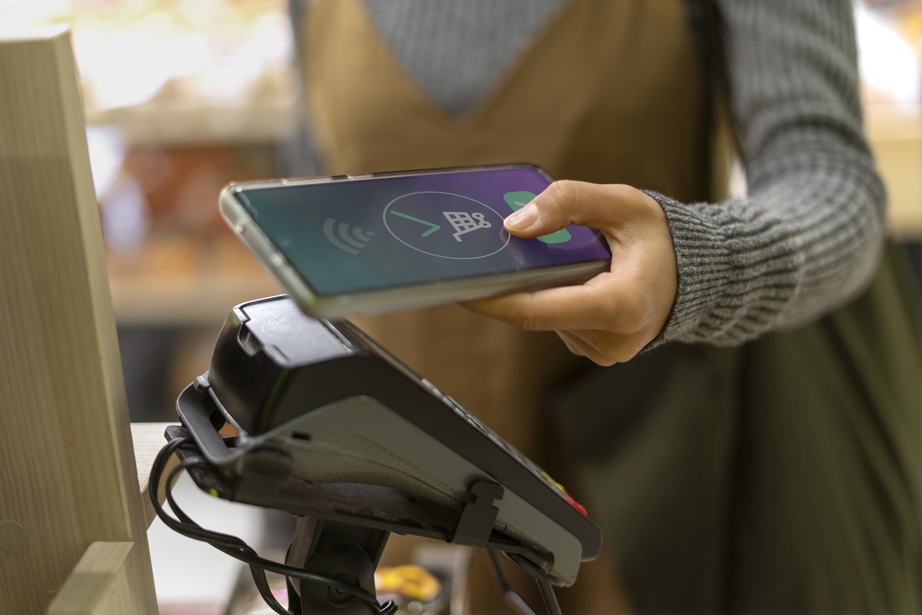 A shopper holds a smartphone over a payment terminal at checkout for a contactless payment, with a shopping cart icon on the phone screen.