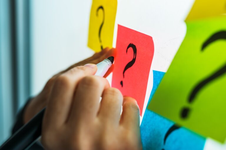 A hand writing question marks on sticky notes: interrogate the requirements before coding
