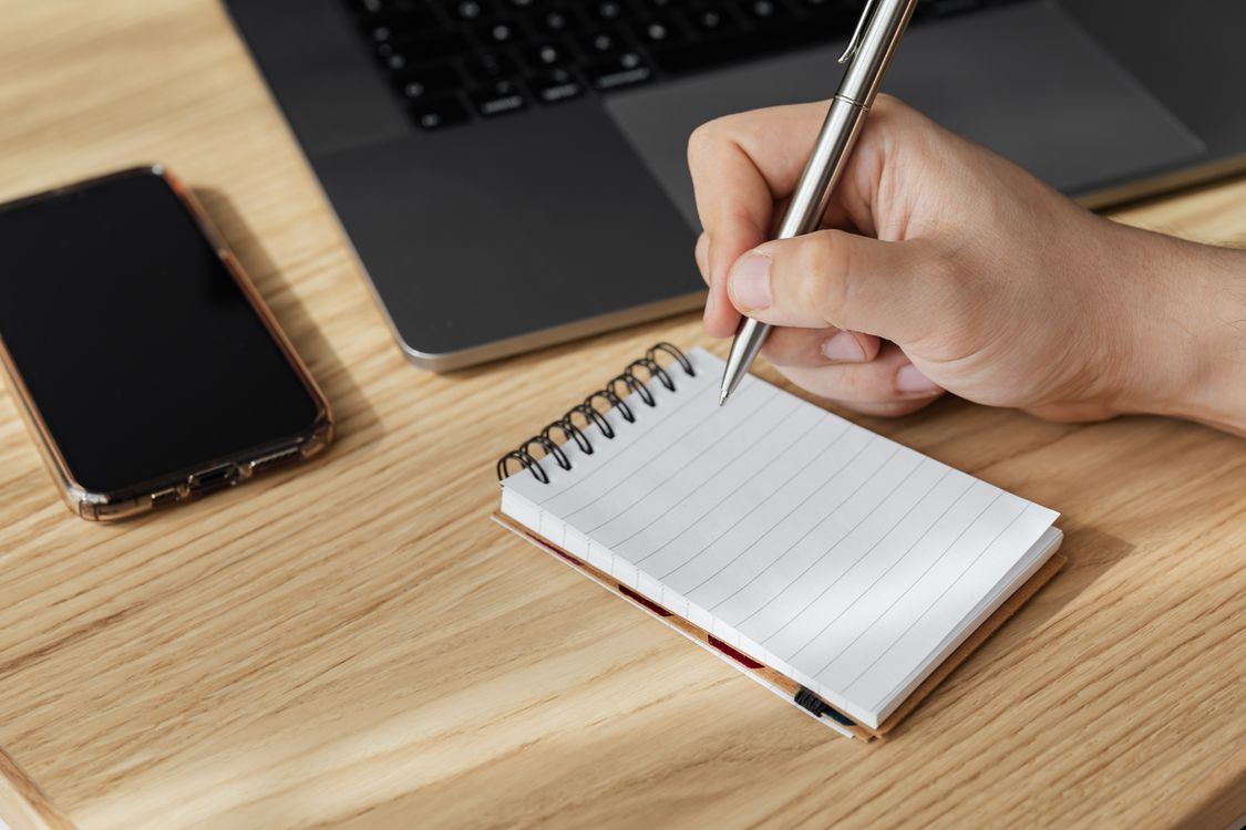 A hand writing in a notebook beside a laptop on a wooden desk